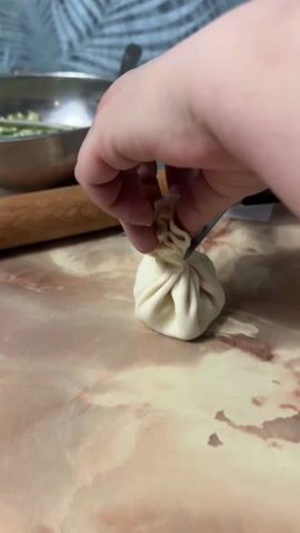 A person uses scissors to cut off the top of a small dough ball, which is formed into a bag shape and filled with a light green mixture. The person's hands are visible, and they are using the scissors to cut the top of the dough ball off. In the background is a rolling pin, a metal bowl with the green mixture, and part of a tropical pattern wallpaper.