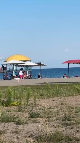 The video shows a beach scene on a sunny day. In the foreground, there is a patch of dry grass and sand. Further back, along the shoreline, there are two covered seating areas, one with a yellow roof and another with a red roof. Several people are visible sitting or lying under these shelters and on the beach. A person wearing a white top and red shorts is walking along the beach path between the grass and the water. The sea is calm and blue under a clear sky.