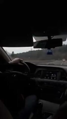 The video shows the interior of a car, likely a Hyundai Sonata, as it drives on a highway. The driver's hands are visible on the steering wheel. The dashboard is visible, showing the car's instruments and infotainment system. Outside the car, the scenery includes trees, an overcast sky, and other vehicles on the road. The overall lighting is somewhat dim. The camera appears to be handheld and positioned from the passenger seat or the back. The audio includes voices speaking, likely having a conversation.
