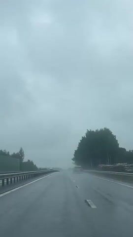 A first-person perspective video shows a car driving on a wet highway during a cloudy day. The road is slick with rain, and visible water spray is coming off the tires of cars ahead. Trees line the right side of the highway, and a metal guardrail is on the left. Other vehicles, including cars and a truck, are also traveling on the highway. The sky is overcast and gray, indicating poor visibility.
