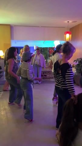 A group of about a dozen women are dancing in a room to music being performed by a man dressed as Elvis Presley. The women are of various ages and dressed casually in pants, tops and some are wearing stripes. The room is decorated in bright colors with plants and modern lighting fixtures. Elvis is sitting on a small stage wearing a white jumpsuit and singing into a microphone. The stage is lit with blue and green lights and has an American flag behind him. Some of the women are watching him and dancing in place.