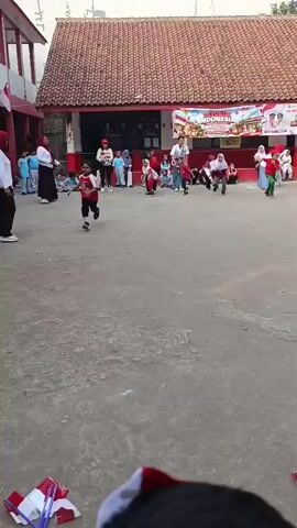 A group of children are running across a paved outdoor area. The children are wearing red and white clothing, and some are wearing hijabs. There is a building with a red roof in the background. A banner is displayed on the building that says "INDONESIA" and appears to be related to a celebration. Some adults are standing around the area, watching the children. There are small Indonesian flags on poles scattered on the ground in the foreground.