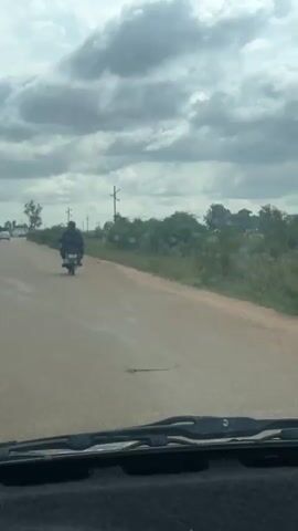 The video shows a view from inside a vehicle looking out at a rural road. The road is paved with a light-colored surface and has a slight curve. On the left side of the road, there are some trees and a few cars in the distance. On the right side of the road, there is a dense line of green bushes and some utility poles. The sky is overcast with dramatic, grey clouds. In the middle of the road, a single person on a motorcycle is riding away from the camera, wearing dark clothing. The vehicle's windshield wipers are visible at the bottom of the frame. The overall impression is of a journey through a somewhat undeveloped, cloudy landscape.