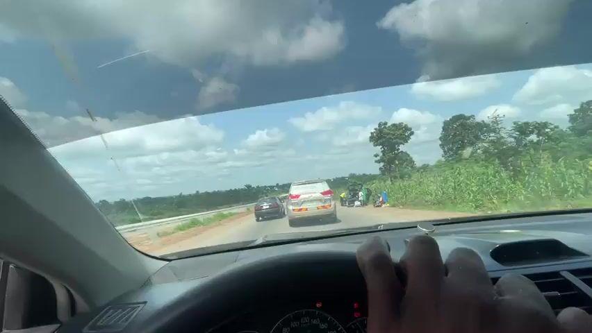 This media file captures a view from inside a vehicle driving on a road in Aciol, Nigeria. The scene is set during the daytime, under a bright sky with scattered white clouds. Lush green vegetation lines the right side of the road, with taller trees visible in the distance. On the left side, there appears to be construction work in progress with exposed earth and barriers.

The primary activity depicted is driving. Several vehicles are visible on the road. In the foreground, the dashboard of the car from which the footage is shot is clearly visible. Ahead, a white SUV is driving in the same direction, followed by other cars and a large red truck. The road itself is paved, with lane markings.

On the side of the road, near the white SUV, there are a few people and some items that appear to be bags or belongings. One person is seen wearing a blue and red garment, possibly a flag or clothing. Another person is seen leaning against the side of the vehicle. The presence of these individuals and their belongings suggests a possible stop or brief pause on the journey.

The overall impression is a journey through a rural or semi-rural area of Nigeria, with active road travel and roadside activities. The weather appears fair and sunny, contributing to good visibility. There is no visible text in the media file.