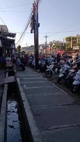 The video shows a street scene from a low angle, looking up towards a bright sky. On the left, there are several stalls or small shops with people standing and sitting around. A narrow, dirty drainage ditch runs along the bottom of the frame. A gray concrete sidewalk is visible, leading towards a large collection of parked motorcycles. The motorcycles are mostly scooters, of various colors, and fill a significant portion of the right side of the frame. Behind the motorcycles, a street is lined with trees and buildings. Red and white flags, possibly national flags, are prominently displayed along the buildings and trees, suggesting a festive or celebratory occasion. A tall utility pole with numerous wires runs vertically through the center of the frame, obscuring some of the background. The lighting suggests it is either early morning or late afternoon due to the long shadows and the quality of the light. The overall impression is a busy street, possibly a marketplace or event space, with a large number of people and motorcycles.