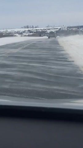 From a vehicle perspective, the video shows a car traveling down a snowy road. The road appears mostly clear, with patches of ice and snow. There is another car ahead on the same road. On either side of the road is snow covered terrain. In the background, a distant village can be seen. The sky is overcast.
