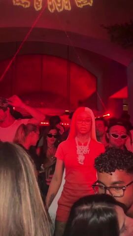 A crowded night event takes place under an archway, with a light display projecting "SLIME" in cartoon letters. A woman in a bright orange t-shirt and shorts, wearing glasses and a large necklace, stands out in the crowd. The crowd is diverse, with various clothing styles and sunglasses visible. The event is lit with red and blue lights, with red lasers crisscrossing the area. The woman in orange stands still as the crowd moves around her, giving a sense of a staged or planned entrance.