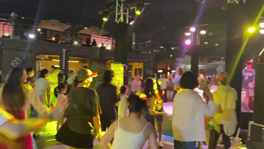 At night, a group of people are dancing under bright stage lights in an outdoor venue. The crowd appears to be engaged in a choreographed dance session, with many individuals moving their arms in unison and following the lead of a person on stage. The lighting is dynamic, shifting between yellow and purple hues, illuminating the participants and the surrounding area. The background reveals a multi-level building with balconies and awnings, suggesting a commercial or entertainment space. Some people in the crowd are children, indicating a family-friendly atmosphere. The overall scene depicts an energetic and lively public event.