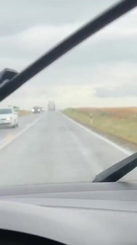 The video shows a view from inside a vehicle driving on a wet road during overcast weather. The windshield wipers are visible in the foreground, and raindrops can be seen on the glass. The road stretches ahead, with two other vehicles, a car and a truck, visible in the distance. The landscape on either side of the road consists of fields.