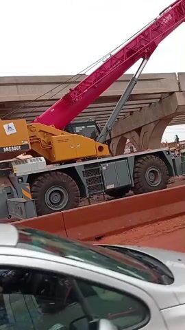 The video shows a construction site with a large pink crane lifting a concrete bridge segment. Below the crane, a silver car is partially visible, and to the right, a yellow auto-rickshaw is also present. The ground is covered in red dirt, and some orange barriers are visible. The sky is overcast.