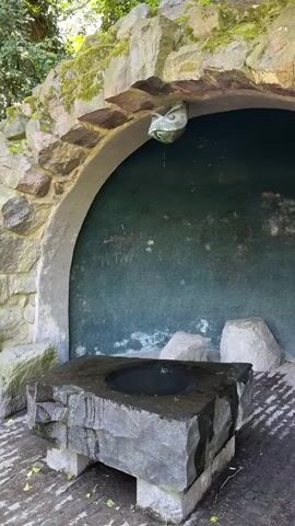 The camera pans up and slightly to the right, showing a stone structure with a curved alcove that serves as a fountain. The fountain is made of rough-hewn stone, and has a square base, on top of which there is a basin filled with water. A small stream of water is trickling into the basin from the top of the alcove. The alcove's backdrop is a textured green surface with some scribbles on it. The structure is built of different sizes and shapes of grey and tan stones. Moss is growing on the structure. There are two large stones next to the fountain's base. The ground is paved with grey stones, and leaves are scattered on the ground. Trees are visible in the background.