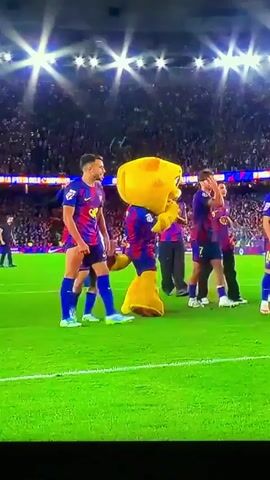 A video depicts the mascot of FC Barcelona, a yellow, smiling cartoon cat named Cati, walking with the team on a soccer field, amid bright stadium lights. The soccer team players are wearing the FC Barcelona uniform, which is blue and red, with yellow lettering. They are clapping and raising their arms to the crowd. The players are walking on green turf. The field is lined with other stadium lights, and a crowd in the background is barely visible. The camera is still.