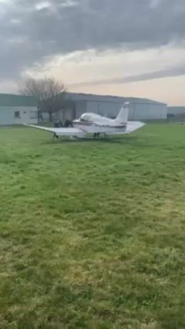 A white and red single-engine airplane sits stationary in a grassy field, with two people standing beside its wing. The plane has the registration "F-BRXE" painted on it. Behind the plane is a large, silver-colored hangar. The sky is overcast with a slightly dull lighting. A small building with green trim is visible to the left of the plane, along with a bare tree. The grass is green and neatly kept. The overall setting seems to be a small airfield or aviation club.