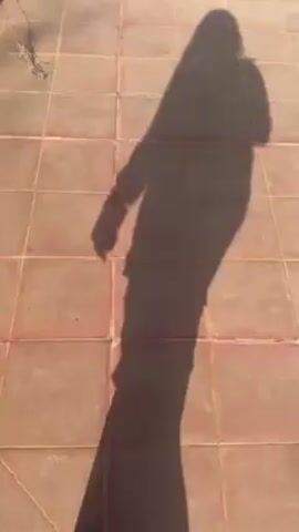 The video shows the shadow of a person standing on a tiled patio that leads to a grassy area with stepping stones. The shadow moves across the tiles and onto the grass. There is a potted plant visible at the edge of the patio. The grass appears dry and patchy. The sunlight is casting a clear shadow, suggesting a sunny day.