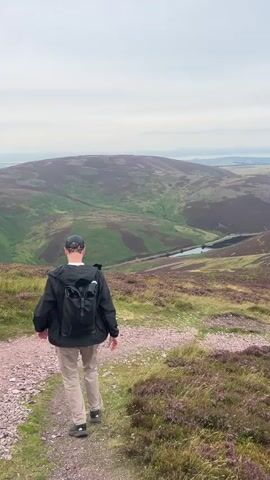 A person wearing a dark jacket, light-colored pants, and a black baseball cap walks along a rocky path on a hillside. They have a black backpack on. The path is surrounded by grass and purple heather. In the distance, there are rolling hills covered in green and purple vegetation. A small body of water is visible in a valley between the hills. The sky is overcast and hazy.
