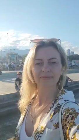 The video is a selfie taken by a fair-skinned woman with blonde hair, wearing a white and gold patterned open shirt over a white top, and a necklace. She is standing outdoors during the day. In the background, a Ferris wheel and various buildings are visible. Other people are present in the background, some sitting and others standing. The overall environment appears to be a promenade or public space, possibly near a body of water, given the presence of a pier-like structure in the last frame. The lighting suggests it is either morning or late afternoon.