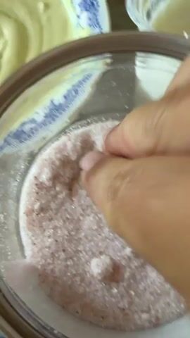 A close-up shot shows a hand reaching into a jar of pinkish salt. The fingers pinch a small amount of salt, which is then sprinkled into a bowl of creamy yellow sauce. The bowl has a blue and white patterned rim. In the background, there are other bowls, one containing a darker, saucy mixture. The lighting is natural, and the focus is on the action of adding salt to the sauce.