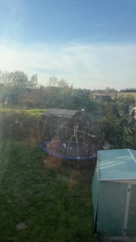 The video shows a view from a window looking out at a backyard. The backyard has a trampoline, a shed, and some overgrown vegetation. There is a fence separating the backyard from the neighboring property. The sky is blue with some clouds. There are water spots on the window. The sun is shining and casting shadows in the backyard.