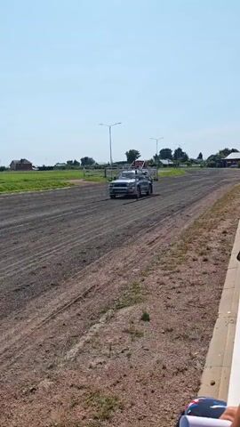 A dark-colored SUV is driving down a dirt track under a clear blue sky. The SUV has a cage-like structure attached to its rear. The vehicle is moving away from the camera. There are buildings and trees in the background, as well as light poles and flags. The ground is dusty and appears to be a race track or similar venue.