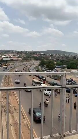 The video shows a busy highway in a city from the perspective of a pedestrian overpass. Cars, vans, trucks, and motorcycles drive on both sides of the highway. A large group of people is congregated on the right side of the road. Some are walking, while others appear to be standing or interacting with vendors. Beyond the highway, there is a view of buildings and hills under a cloudy sky. The overpass has a metal railing. The highway has yellow lines dividing the lanes. The ground on either side of the highway appears dry and somewhat barren.