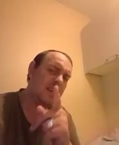 A man with receding hair and a brown t-shirt is looking into the camera. He is in a room with a peach-colored wall and a white cabinet on the right side of the frame. There are some items wrapped in plastic on a surface behind him. The man is speaking and gesturing with his hands.