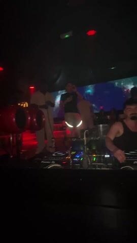The video captures a lively nightclub scene bathed in red lighting. Initially, the focus is on a stage where several people are performing, including a DJ with equipment in front of them and others who appear to be playing percussion instruments and singing. The camera then pans upwards and outwards, revealing a crowded dance floor filled with people of various ages and ethnicities. Many are dressed in dark clothing, and they are dancing and socializing. A disco ball hangs from the ceiling, adding to the festive atmosphere. The venue has multiple levels, indicated by the red-lit railings in the background.