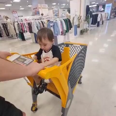 A child sits in a yellow shopping cart in a store. The child is wearing a black and white shirt. The cart is being pushed by an unseen person whose arm and hand are visible in the lower left corner of the frame. The background shows racks of clothing and displays in a retail environment. The video is shaky, suggesting movement.