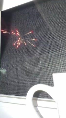 A white mug is positioned on a windowsill. Beyond the mug, a dark window screen is visible, through which a nighttime sky can be seen. Fireworks briefly explode in the sky at different intervals. The fireworks appear as bursts of light, with some emitting red and yellow sparks. The screen obscures some of the detail of the fireworks and the sky.