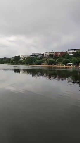 The video captures a serene lake scene in Angelovo, Russia, under an overcast sky. The camera pans across the water, reflecting the grey clouds above and the houses lining the distant shore.

The subjects in the scene are primarily the natural elements of the lake and its surroundings, along with several houses and trees situated on a hill overlooking the water. There are no visible people or specific objects of focus, emphasizing the tranquility of the landscape. The setting is a body of water with a developed shoreline, suggesting a residential or recreational area.

The activity taking place is a slow pan of the camera, moving from left to right across the lake, revealing the different parts of the landscape. A notable detail is a powerful water fountain shooting a tall stream of water high into the air, creating a dynamic element against the otherwise still scenery. The time of day appears to be late afternoon or early evening, given the soft, diffused light. The weather is cloudy, contributing to the subdued atmosphere. There is no visible text in the video. The overall emotion conveyed is one of peacefulness and quietude.