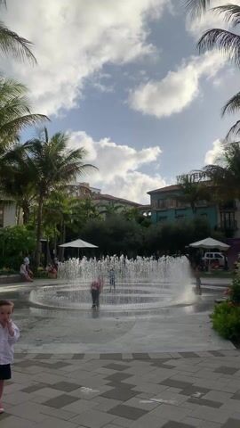 The video shows a central plaza area with a large circular fountain as its focal point, with numerous water jets spraying upwards, creating a visually engaging display. Several children are playing inside the fountain. Surrounding the fountain are palm trees, providing a tropical ambiance, and there are buildings with reddish-brown roofs in the background, one of which has the words "WINWOOD" on it. The sky is partly cloudy with patches of blue. A young boy in a white shirt and dark shorts is visible in the foreground on the left, and several people are seated around the area under white umbrellas and on benches, giving a relaxed and communal atmosphere.