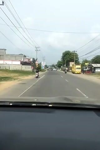 The video shows a view from inside a car driving on a road. The scene is an outdoor, daytime setting, likely in a semi-urban or rural area, given the presence of houses, shops, and utility poles with many overhead wires. The weather appears to be overcast or hazy.

The road is paved and has at least two lanes. There is traffic on the road, including several motorcycles, cars, and a yellow truck. On the left side of the road, there are buildings, some with signage, and vegetation. On the right side, there are more buildings, trees, and more utility poles.

A person is visible riding a white scooter on the left side of the road in the foreground. Further down the road, other vehicles and people are moving. The overall activity is that of traveling by car through a populated area.

There is some visible text on signs, although it is not fully legible. One sign on the left appears to have "76" on it, possibly a brand. Another sign further down the road has a blue and white design. A sign on the right side shows a green background with white text.

The time of day is daytime, suggested by the natural light, but the sky is not clear blue, indicating potential haze or clouds. The location cues of utility poles with numerous wires, building styles, and vehicle types suggest a Southeast Asian context, and the prompt specifies Payameuneng, Indonesia. There are no overt emotional cues expressed, as the view is from a vehicle.