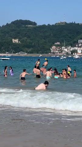 A group of people, primarily children and teenagers, are playing in the ocean near a beach. Some are standing in the water, while others are swimming or playing with a yellow ball. In the background, there is a coastline with buildings and trees, and several boats are anchored in the water. The sky is clear and blue, indicating a sunny day. The waves are small to moderate, washing onto the sandy shore in the foreground.