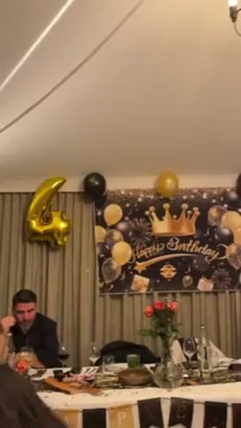 The video captures a birthday celebration. Guests are seated around a decorated table, with a "Happy Birthday" backdrop adorned with black and gold balloons. A large gold number "40" balloon floats nearby, indicating the age being celebrated. The table is set with drinks, flowers, and celebratory decorations. Guests are interacting, some are clapping and some are talking. A radiant heater is visible on the ceiling.