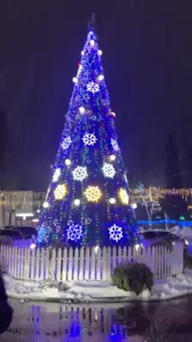 The video shows a nighttime outdoor scene of a Christmas display in the snow. The camera pans from a large Christmas tree decorated with blue and yellow lights, and snowflake ornaments, to a street lined with snow, lit by blue lights and garland with pendant lights.  It is snowing. The area appears to be decorated for the holidays.