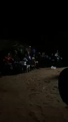 The video shows a group of people gathered outdoors at night. They are sitting on chairs or standing, and there appears to be a small table nearby. The area is dimly lit, with a light source illuminating the group, but details are difficult to make out due to the darkness. The ground is sandy, and there are some natural elements like branches or vegetation visible in the background. People are talking and interacting. The recording seems to be taken from within the crowd, moving and focusing on different areas.
