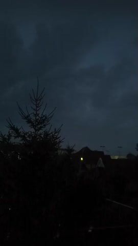 A dark, cloudy sky looms over a shadowy landscape. A lone pine tree stands in the foreground, silhouetted against the gloom. In the background, a few houses with lit windows are visible, suggesting a residential area. The overall atmosphere is dim and somber, with the heavy clouds hinting at impending rain or a storm.