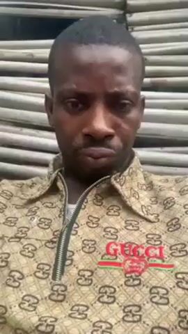 The video shows a man looking directly at the camera and speaking. He has dark skin and a closely shaved haircut. He is wearing a patterned shirt with a "Gucci" logo on the front. The background appears to be a wall constructed of light-colored wooden slats. He is speaking but his words are not clear.