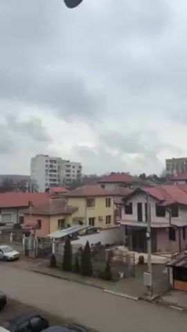 The video shows a residential neighborhood on an overcast day. Several houses with red-tiled roofs are visible, along with taller apartment buildings in the background. A street runs in front of the houses, with cars parked on the side. Trees are interspersed throughout the neighborhood, some bare of leaves.