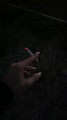 The video opens with a close-up of a hand holding a lit cigarette. The background is dark, suggesting it is nighttime or a dimly lit area. The hand has long, manicured fingernails with a light-colored polish. The cigarette glows red at the tip. The scene then transitions to a wider shot of a dark landscape with a vibrant sunset. Silhouettes of trees and hills are visible against a gradient sky of deep blue transitioning to orange and red at the horizon. The overall mood is somber and atmospheric.