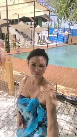 A woman is taking a selfie in front of an outdoor swimming pool. She is wearing a blue sarong over a swimsuit and a necklace. Behind her, the pool is visible with several people swimming. The pool area has a wooden deck, lounge chairs, and umbrellas for shade. In the background, there is a building with multiple levels and some greenery. A man in swim trunks is standing near the edge of the pool. The sun is shining, creating bright light and shadows.