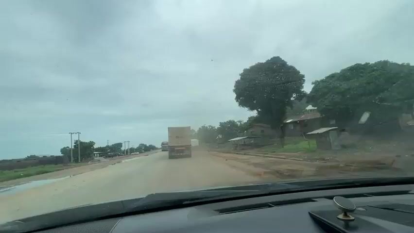 This is a first-person perspective video, likely filmed from the dashboard of a vehicle traveling on a road. The scene depicts a highway in Koton Karifi, Nigeria, under an overcast sky suggesting a cloudy or possibly rainy day.

The primary subjects are several large trucks on the road. In the foreground, the dashboard of the vehicle is visible, including a dashboard mount for a phone. Ahead, a convoy of trucks is moving along the asphalt road. One truck, closer and on the left side of the frame, is a brown articulated lorry with a large, tan-colored tarpaulin-covered cargo. Further ahead, a white box truck is visible, and behind it, another truck with a white tarpaulin. The road appears to be a main thoroughfare, with some signs of construction or wear, and a dirt shoulder.

On the right side of the road, there are some small, simple houses with corrugated metal roofs, indicative of a rural or semi-urban settlement. Lush green trees and foliage are present, especially on the rising terrain behind the houses. On the left side of the road, there are more trees, some vehicles parked or abandoned, and what appear to be large metal tanks, possibly industrial or storage. Power lines are also visible.

The activity is the movement of vehicles along the road, suggesting travel or transportation. The weather is dull, and the overall mood conveyed is that of a typical journey on a Nigerian highway, with the presence of heavy cargo transport being a significant element of the scene. No specific emotions are overtly displayed or implied, beyond the neutral observation of the journey. There is no visible text in the video, except for potential logos or markings on the trucks which are too indistinct to be read.