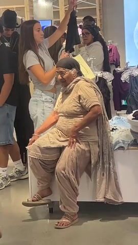 The video shows an elderly woman in a clothing store. She is wearing traditional Indian clothing, a beige salwar kameez with a matching dupatta. She is seated on a white display table and is looking to her left. To her right, a young woman in a grey t-shirt is holding up a black garment. Behind them, other shoppers are visible, including a young man in a black hoodie and another in a black t-shirt. The store appears to be well-lit with racks of clothing in the background. The woman adjusts her scarf and then shifts her weight as if to stand up.