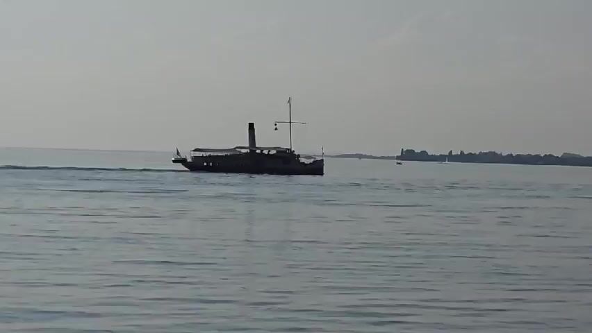 A distant, silhouetted paddle steamer cruises across a wide expanse of water under a hazy sky. The vessel appears to be full of passengers. In the far distance, a faint, urban shoreline is visible, dotted with buildings and a few other small boats. The water is calm with a gentle ripple. The foreground is dominated by a blurred shot of a gravelly shoreline, with the rear wheel of a bicycle partially visible. The overall lighting suggests it is daytime, likely late afternoon or early morning due to the haze.
