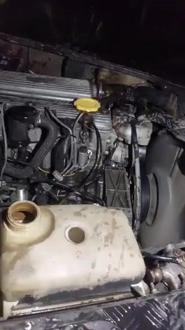 Here's a description of the video:

The video shows the open engine compartment of a vehicle, likely a Land Rover Defender, running. A large, white coolant reservoir is visible in the foreground. The engine is dirty and has some visible components such as hoses, a black air filter, and a yellow cap. The fan is spinning quickly.

A person wearing a gray shirt and a watch on his left wrist appears briefly in the frame. He seems to be doing some work near the engine compartment. 
The video is taken at night, with artificial lighting illuminating the engine. The surrounding area is dark.