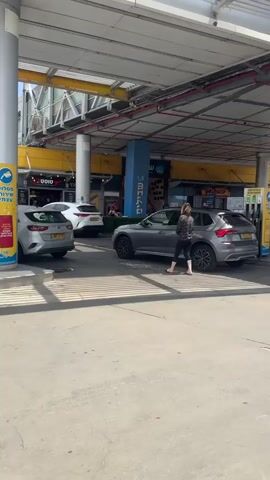 The camera pans across a gas station on a bright day. A woman stands near a gray SUV filling up with gas at a pump under a yellow canopy. Next to the SUV is a white pickup truck.  Other vehicles fill up at pumps farther down under the canopy, including a military-style Jeep and white sedan. A building sits behind the canopy with various businesses.