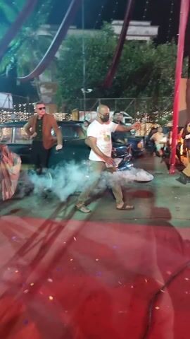 The video shows a busy outdoor event at night, possibly a party or wedding reception. People are dressed in festive attire and mingling under decorative lights and streamers. Some individuals are dancing, while others are seated at tables. A person is standing on a table in the distance. A man is walking while holding a smoke emitter. The atmosphere is lively and celebratory.