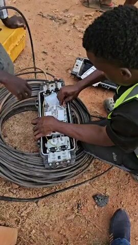 Two men are working on telecommunications equipment outdoors. A long spool of black cable surrounds a large rectangular gray box, which one man is working inside. The box is open and revealing a complex arrangement of wires and components. One man, wearing a black shirt and a bright green reflective vest, is kneeling and carefully manipulating the equipment with his hands. The other man, whose lower body is visible, holds a small white packet. A yellow toolbox sits nearby. The environment is a dirt surface. The weather appears to be sunny.