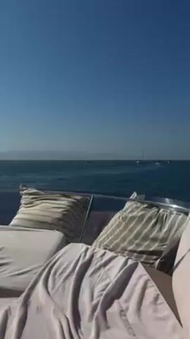 The video shows a view from the back of a boat on a sunny day. The sea is calm and blue. There are a few distant boats visible on the horizon. In the foreground, there are pillows and a blanket on a lounge chair. The sky is clear blue.