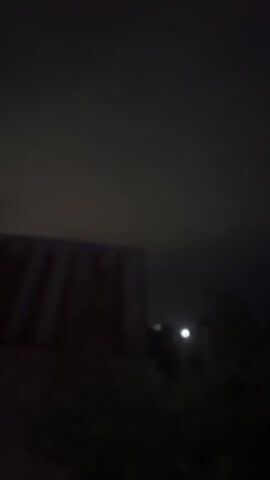 The video shows a dark, nighttime scene. On the left, there is a dark, rectangular structure, possibly a building. In the middle-right, a bright light, potentially the moon, is visible through some dark foliage. The sky is overcast and dark. The scene is still and there are no visible people or animals. The overall impression is one of an outdoor, nocturnal setting, with limited visibility due to darkness and clouds.
