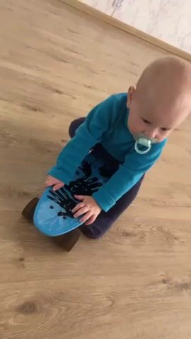 A baby wearing a teal long-sleeved shirt and navy blue pants kneels on a wooden floor, gripping a blue skateboard with black handprints on it. The baby has a pacifier in its mouth. The skateboard's wheels are visible. A crumpled piece of white paper lies on the floor to the right of the skateboard. The background shows a light-colored wall with a subtle pattern and a hint of a wall socket. The camera angle is from above and slightly to the side, looking down at the baby and the skateboard. The video is short, with minimal movement.